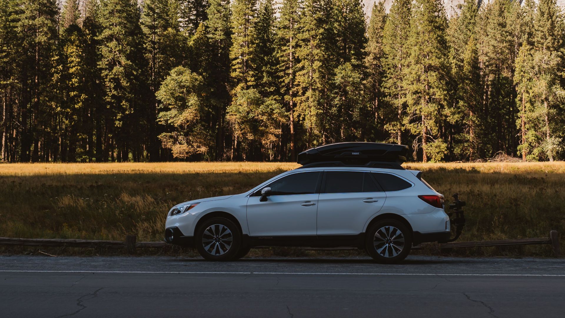 A white SUV parked on a peaceful road beside lush trees and rocky cliffs possibly in Yosemite National Park.