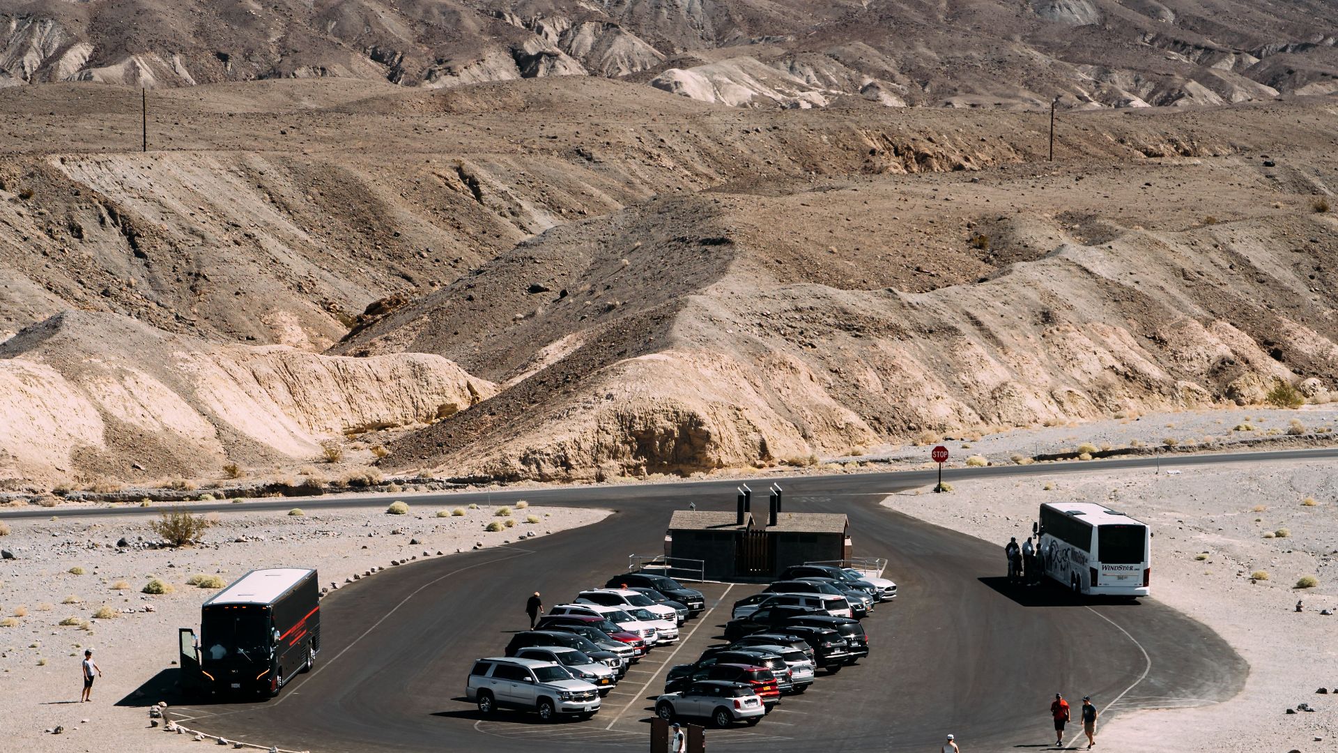 Tourists explore a desert parking area with cars and buses, set against mountain backdrop.