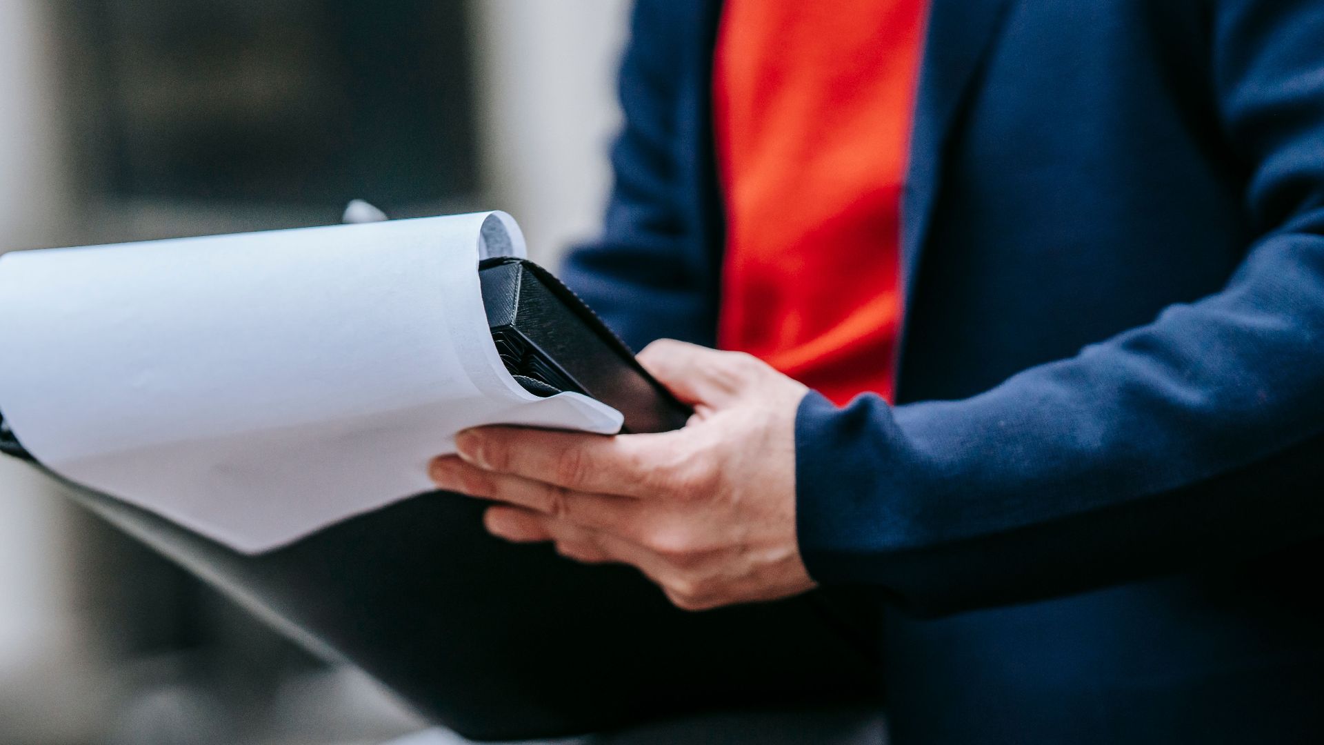 A professional adult outdoors in a suit jacket, focused on reviewing paperwork, highlighting modern business style.