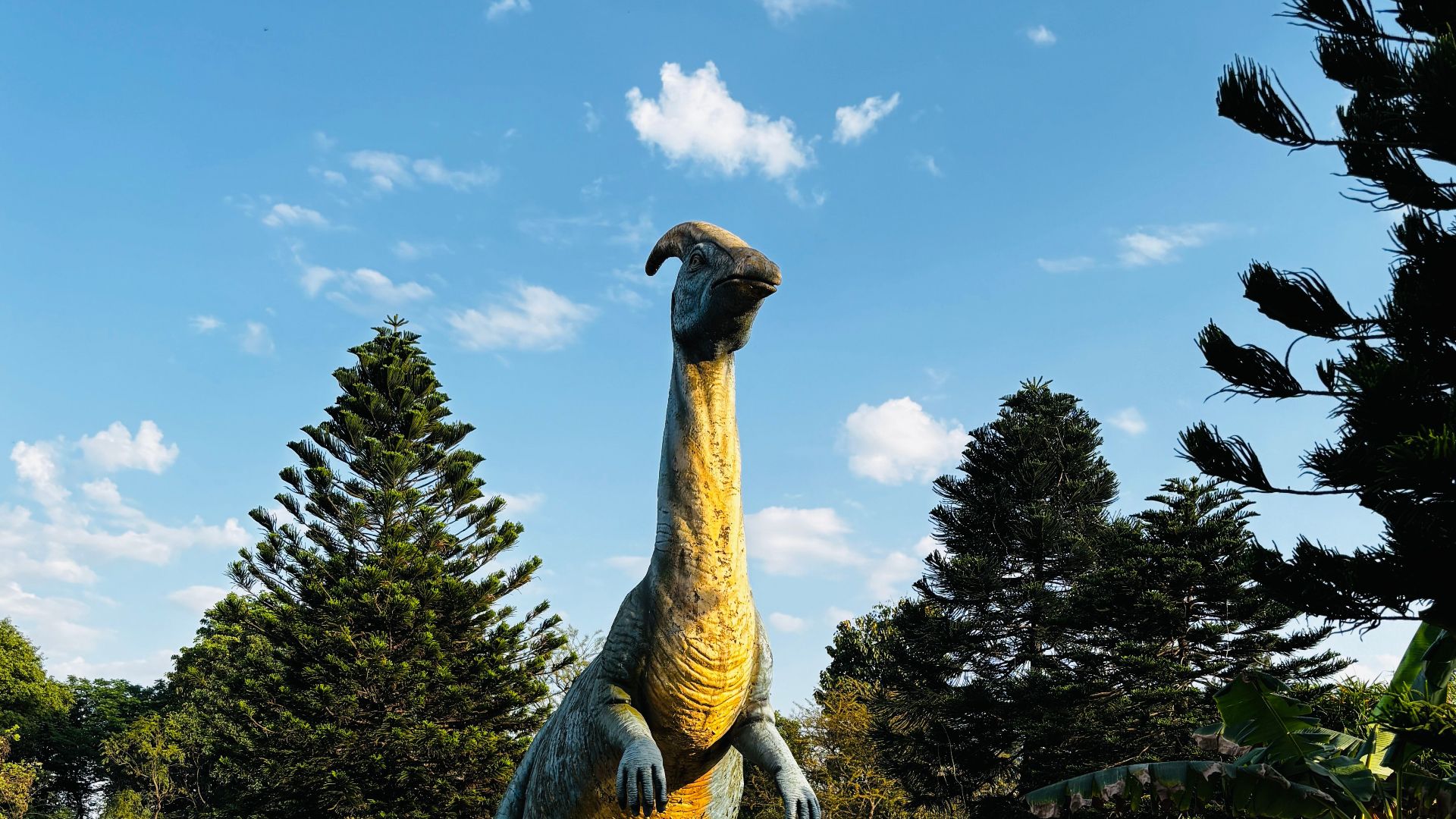 A lifelike dinosaur statue in a landscaped park with blue skies and lush greenery.