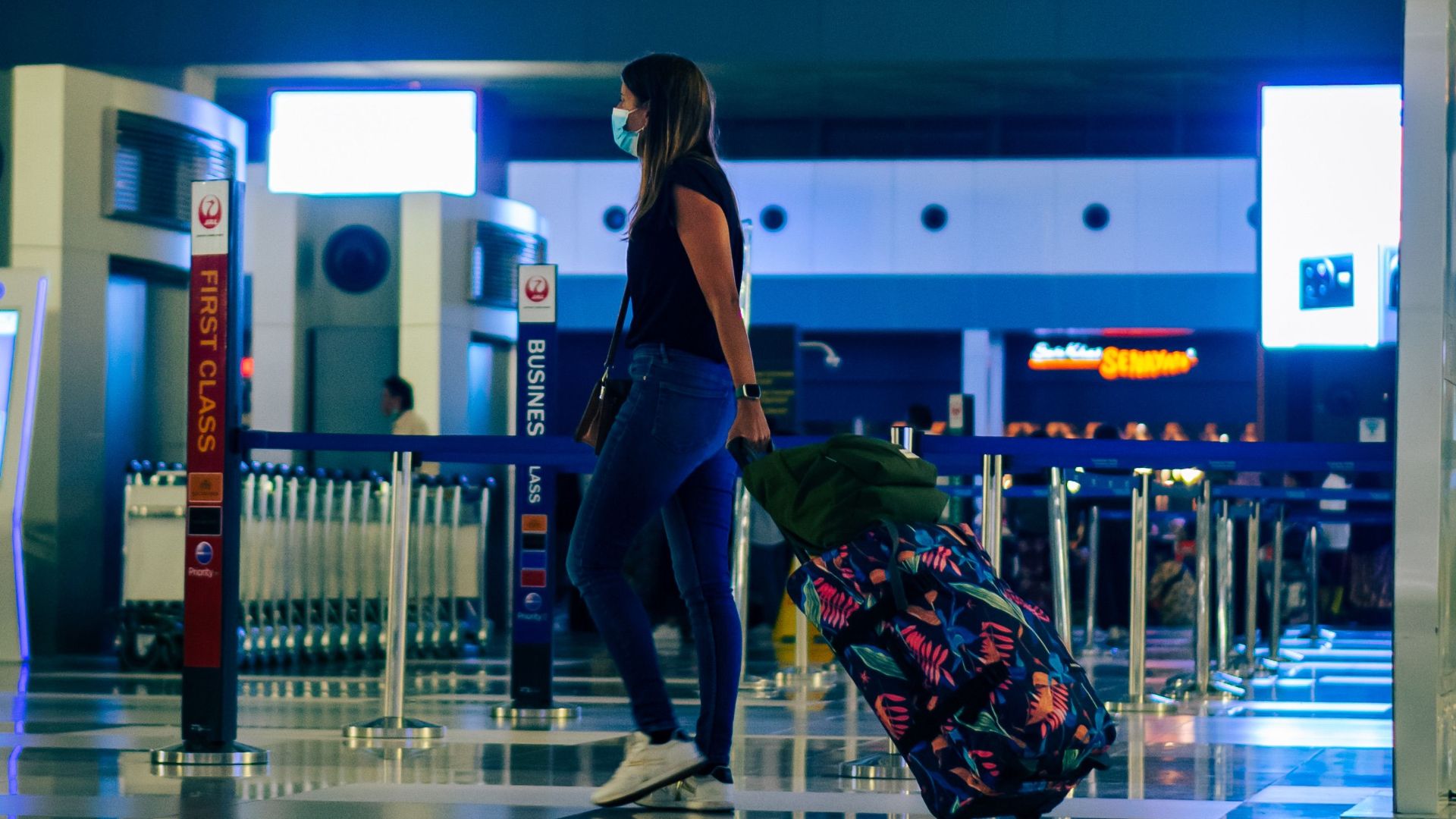 A woman wearing a face mask walks through an airport with luggage, reflecting modern travel trends.