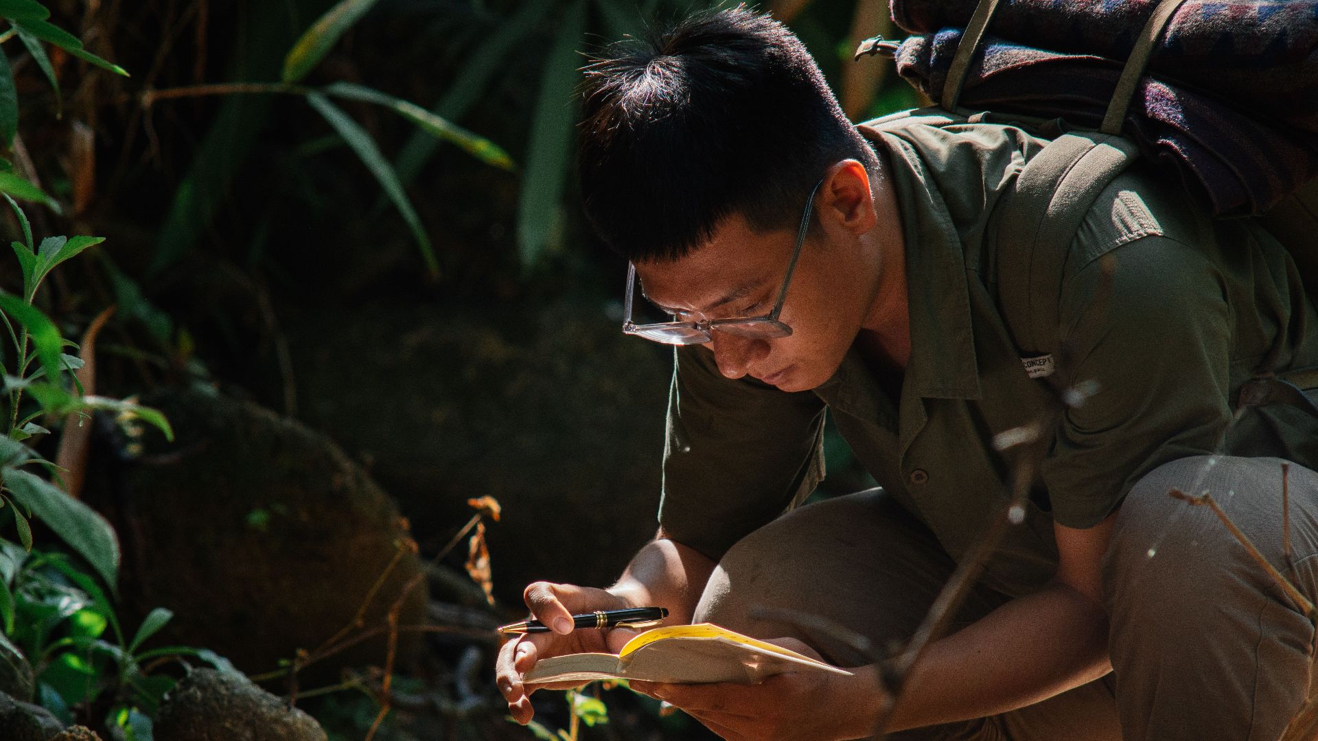 Man crouching outdoors, writing in a notebook surrounded by lush greenery, showcasing a peaceful, contemplative moment.