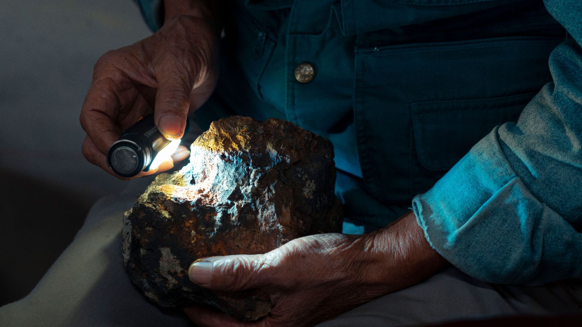 Detailed close-up of hands using a flashlight to examine a geological stone indoors.