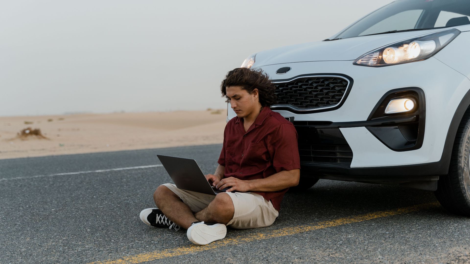 A man using a laptop while sitting on the road beside a parked car in a desert setting.