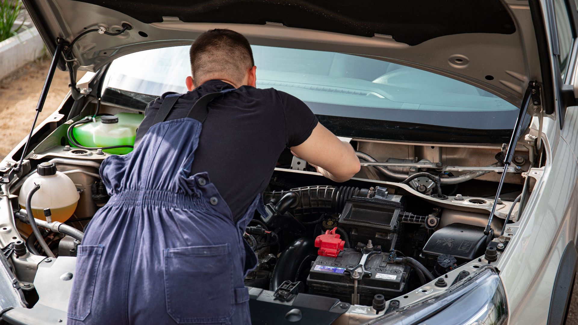 Mechanic in overalls working on a car engine outdoors, showcasing vehicle repair skills.