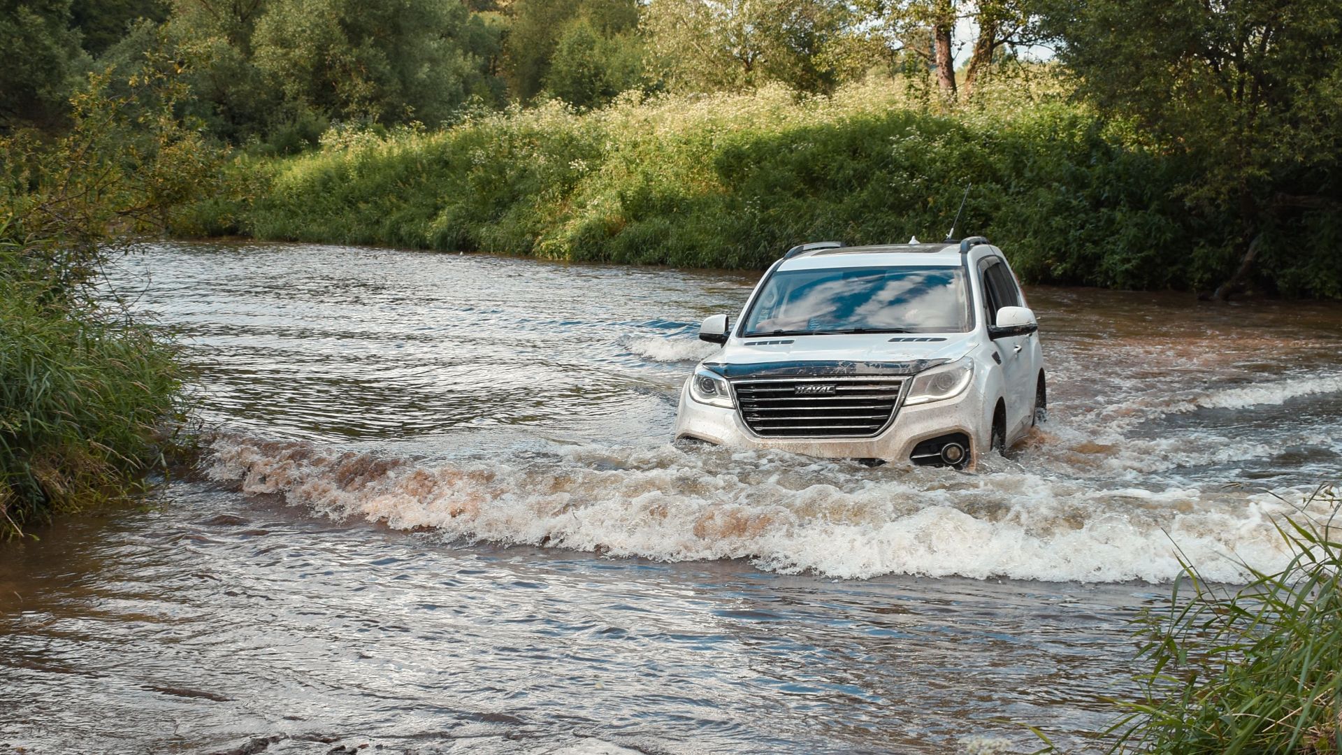 An SUV crosses a shallow river surrounded by lush greenery, showcasing off-road capabilities.