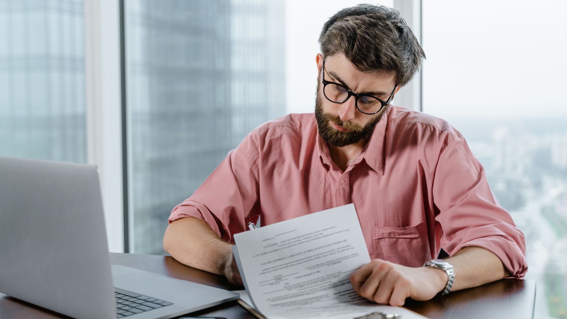 A businessman in glasses examining documents at a desk with a laptop, indoors.