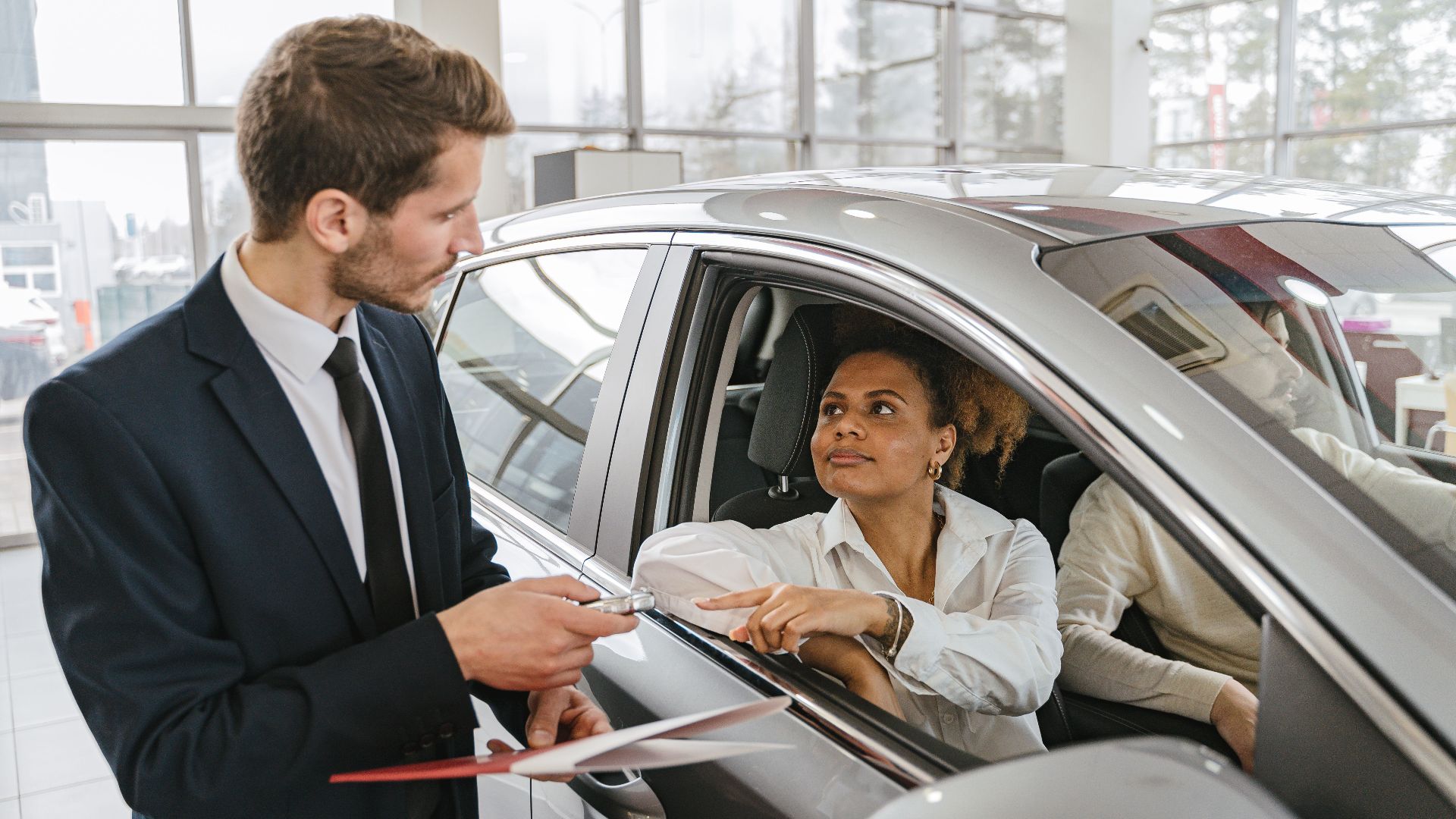 A car dealer hands keys to a woman sitting inside a vehicle in a car showroom.