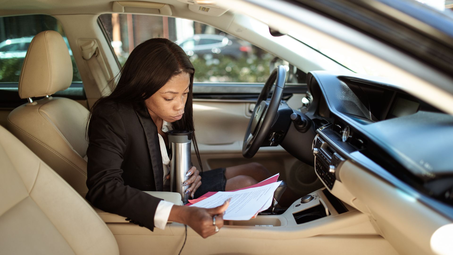 A focused businesswoman reads documents in her car, multitasking with a tumbler.