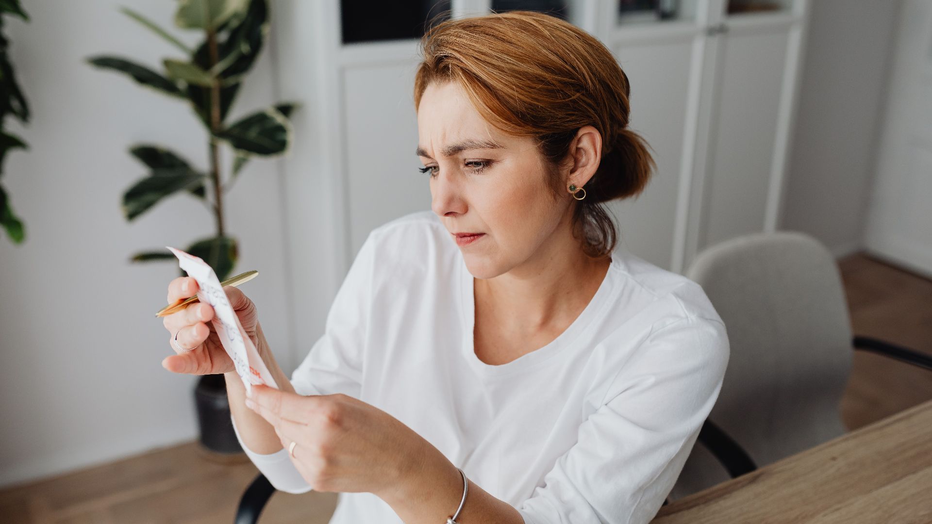 A concerned adult woman examines a receipt while sitting at her office desk, indicating financial scrutiny.