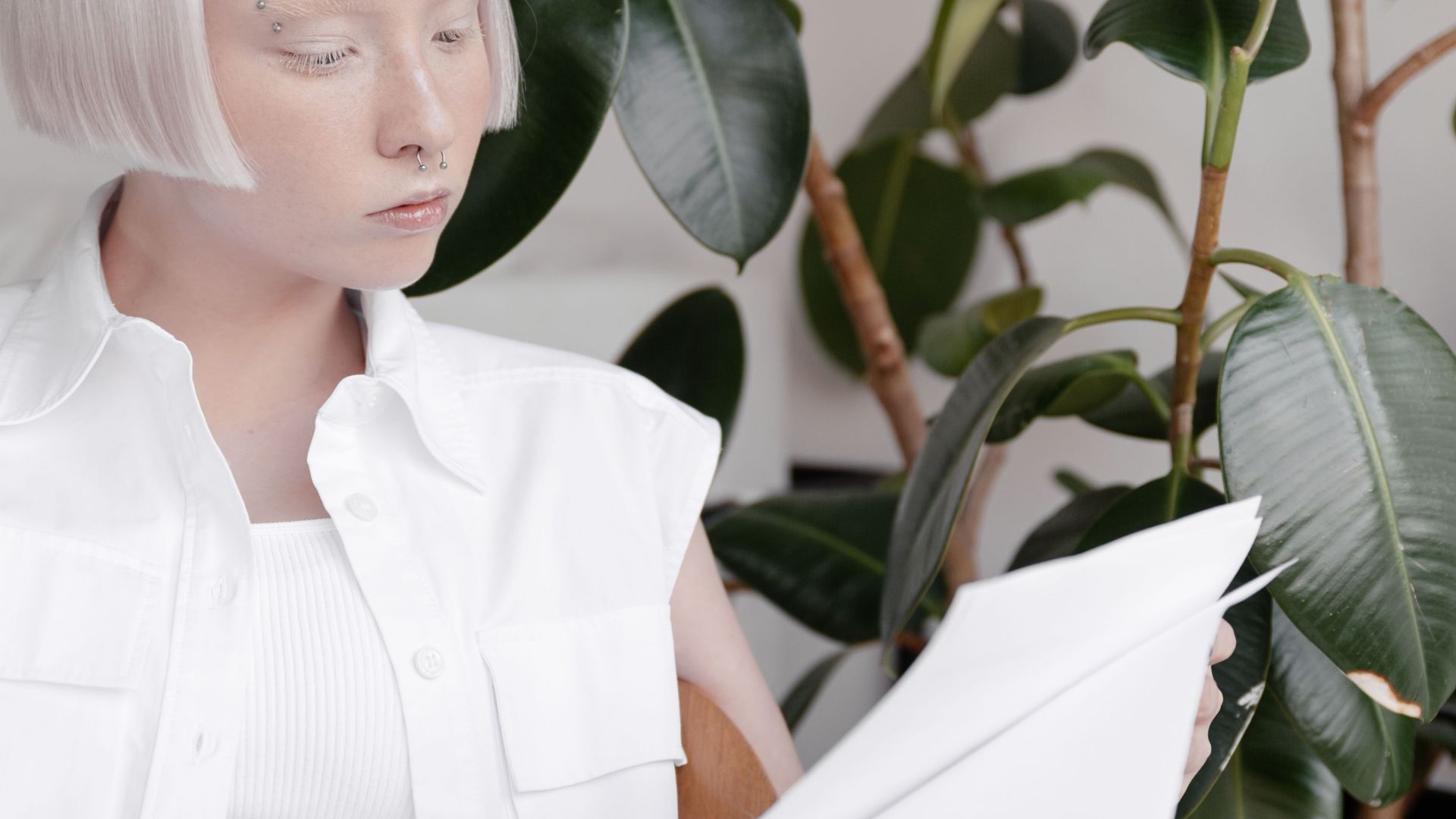 A woman with platinum hair reads a document in a stylish, plant-filled interior.