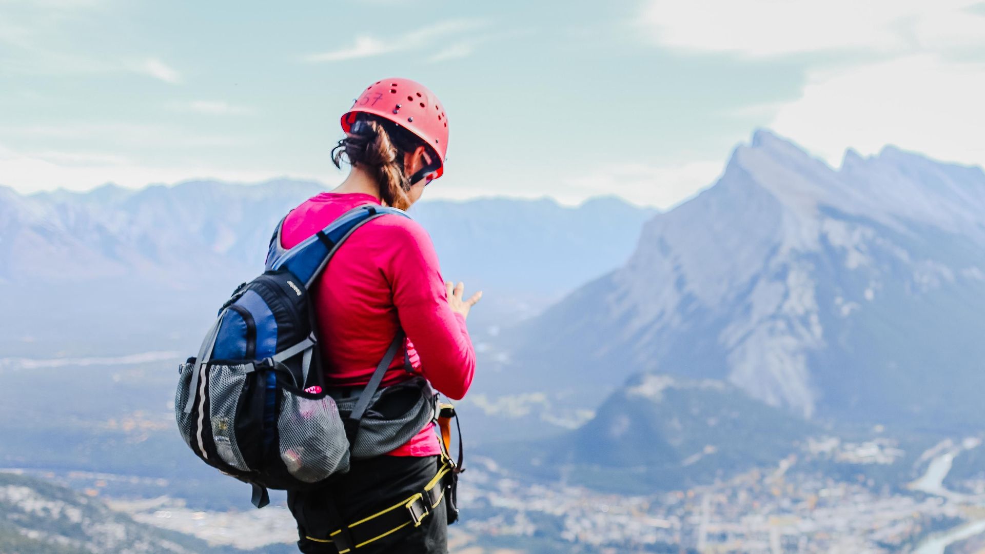 Woman wearing a pink sweater and helmet hiking on a mountain cliff with a scenic view.