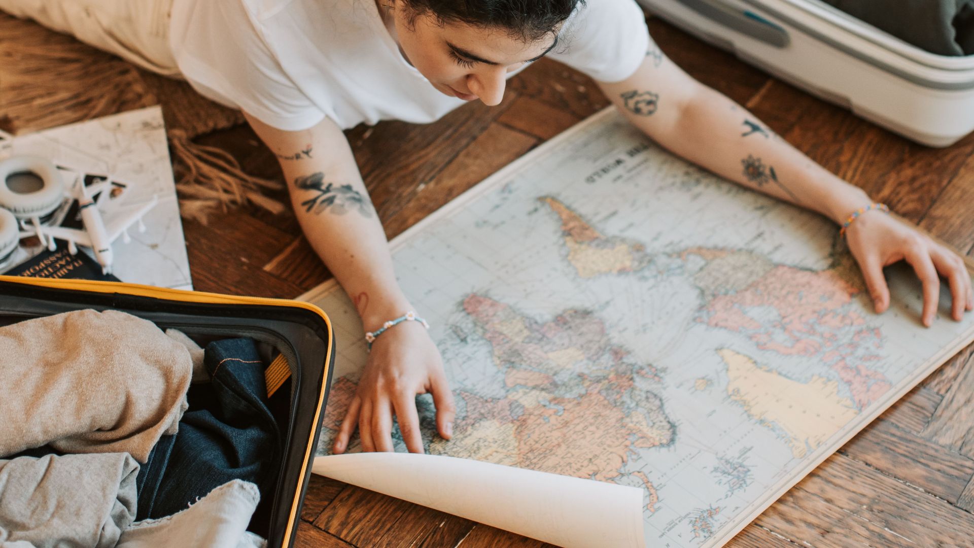 A woman lying on the floor indoors, planning a trip with a map and open suitcase.