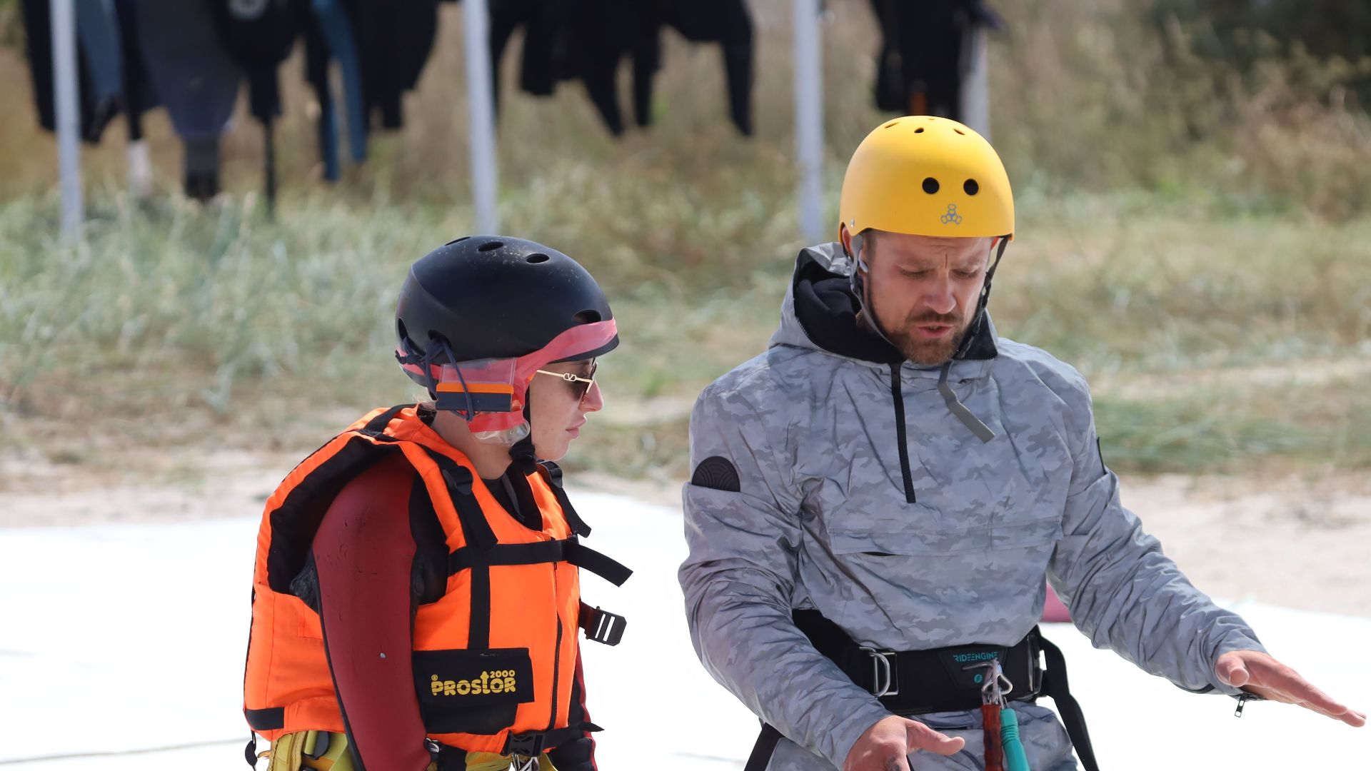 Instructor and student kneeling on beach during outdoor kitesurfing lesson, using safety gear.
