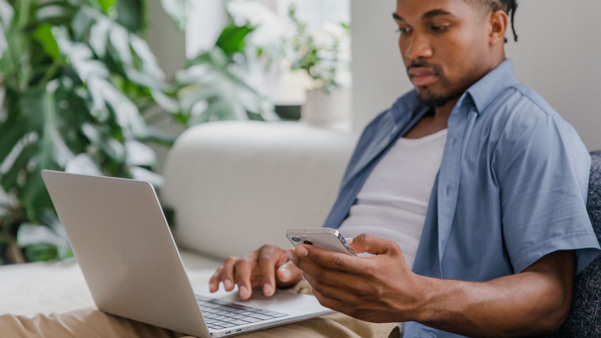 A young man multitasks with a laptop and smartphone on a sofa, dressed casually indoors.