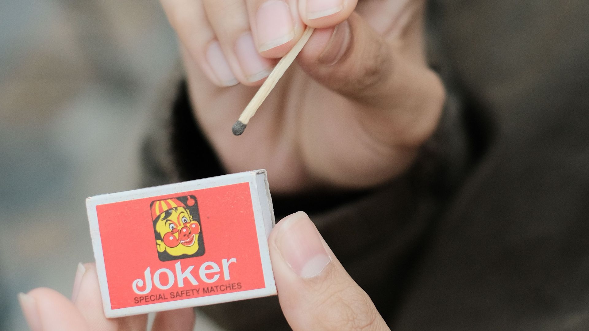 A person holding a matchstick and a matchbox labeled 'Joker' in a close-up shot.