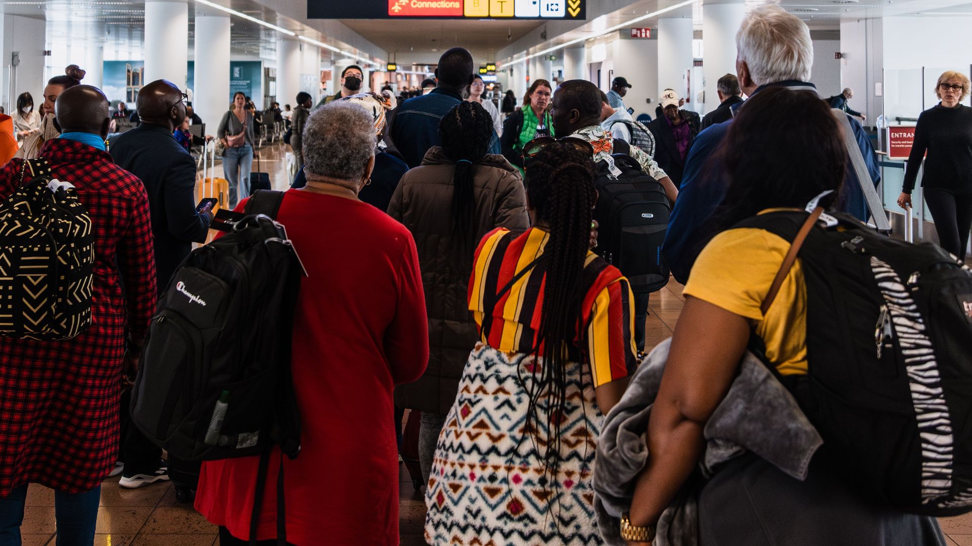Travelers lining up in a busy airport terminal with baggage under flight information screens.
