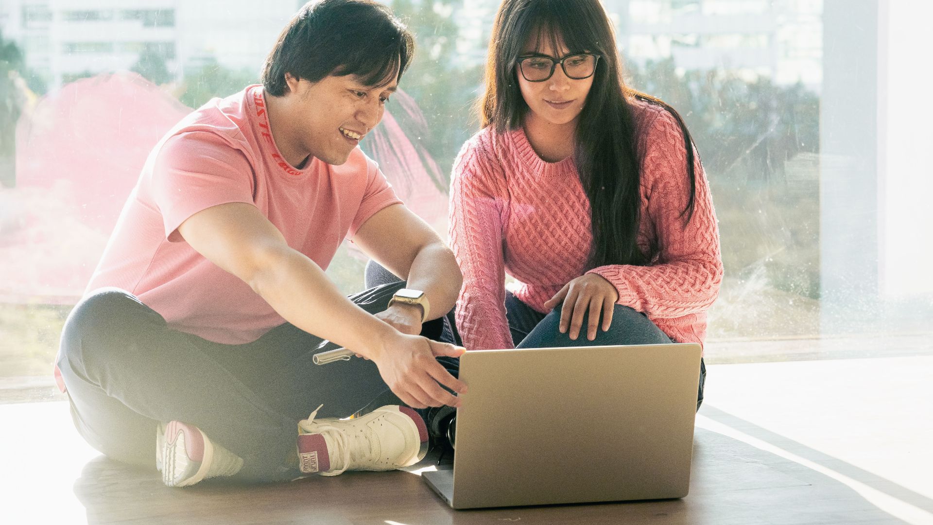 Two young adults in casual attire working together on a laptop by a window.