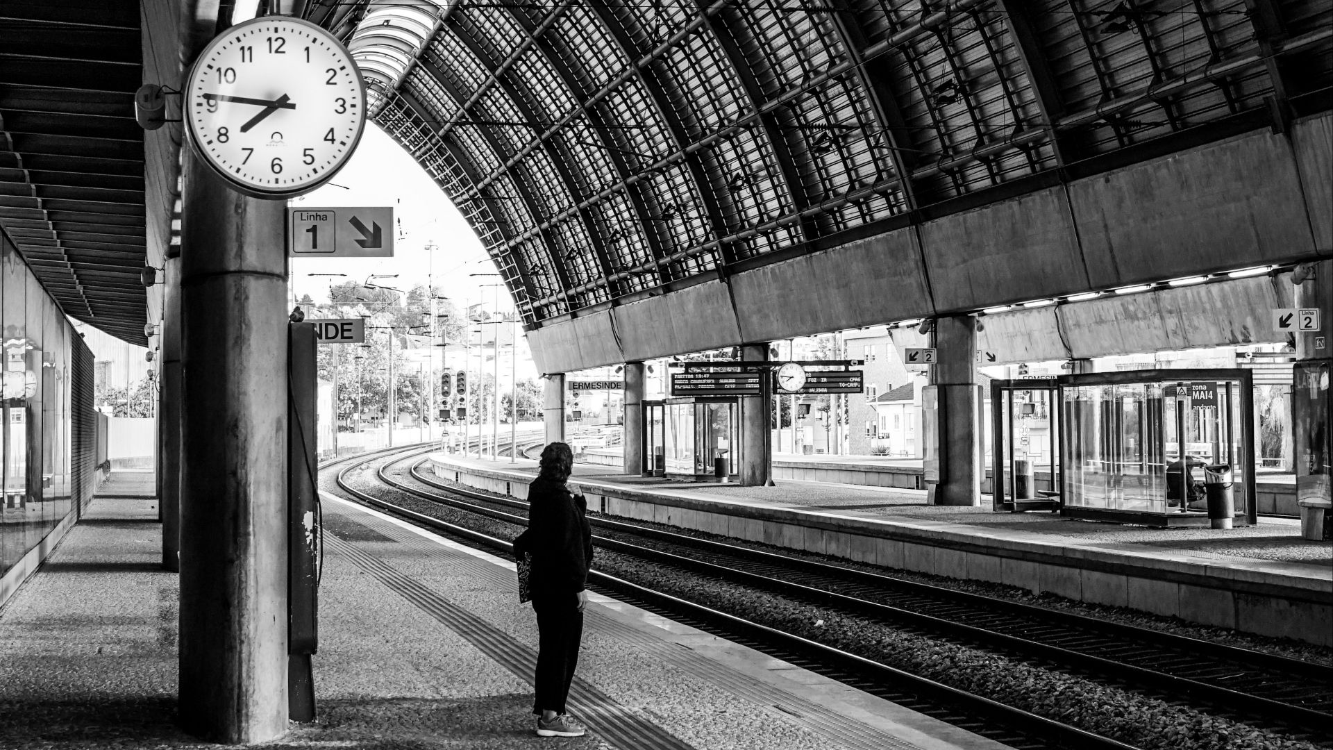 Black and white photo of a person standing in a railway station with a clock overhead.