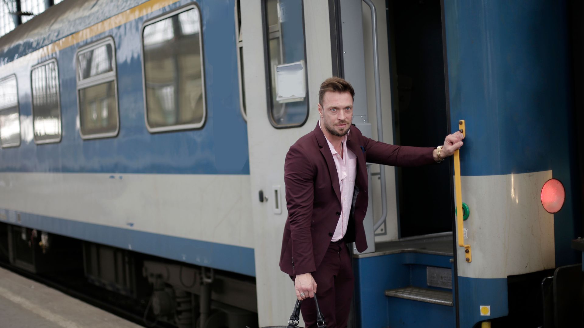 Serious male passenger with travel bag wearing formal outfit standing near train wagon with opened door before entering on railroad station during business trip