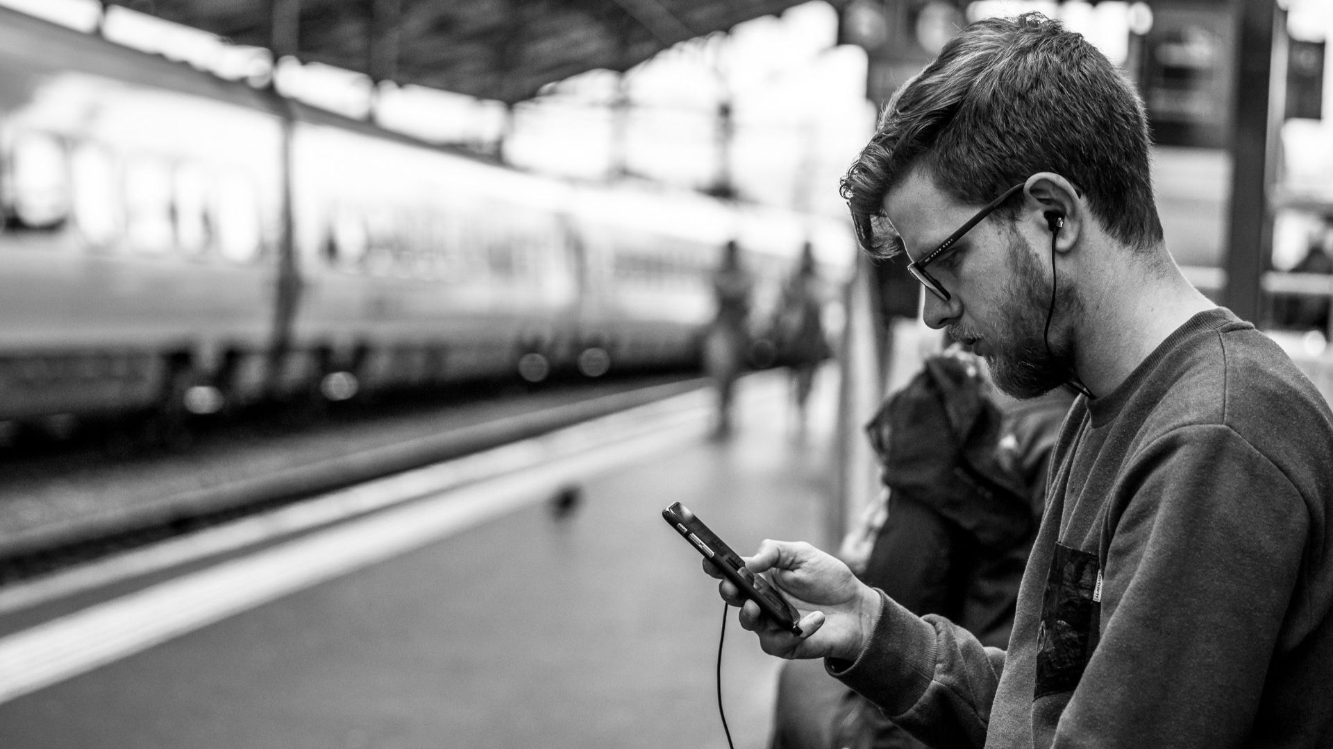 Black and white photo of a man with earphones using a phone at a train station in Lausanne, Switzerland.