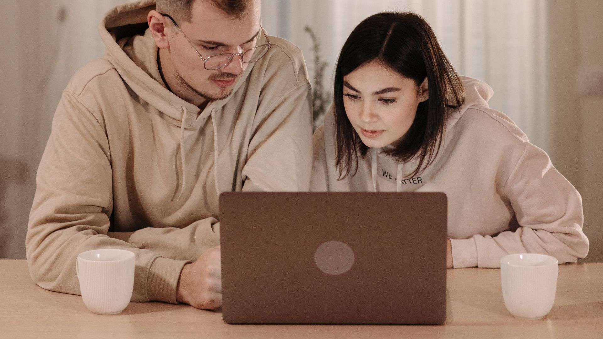 A couple in hoodies working on a laptop in a cozy home setting with warm lighting.