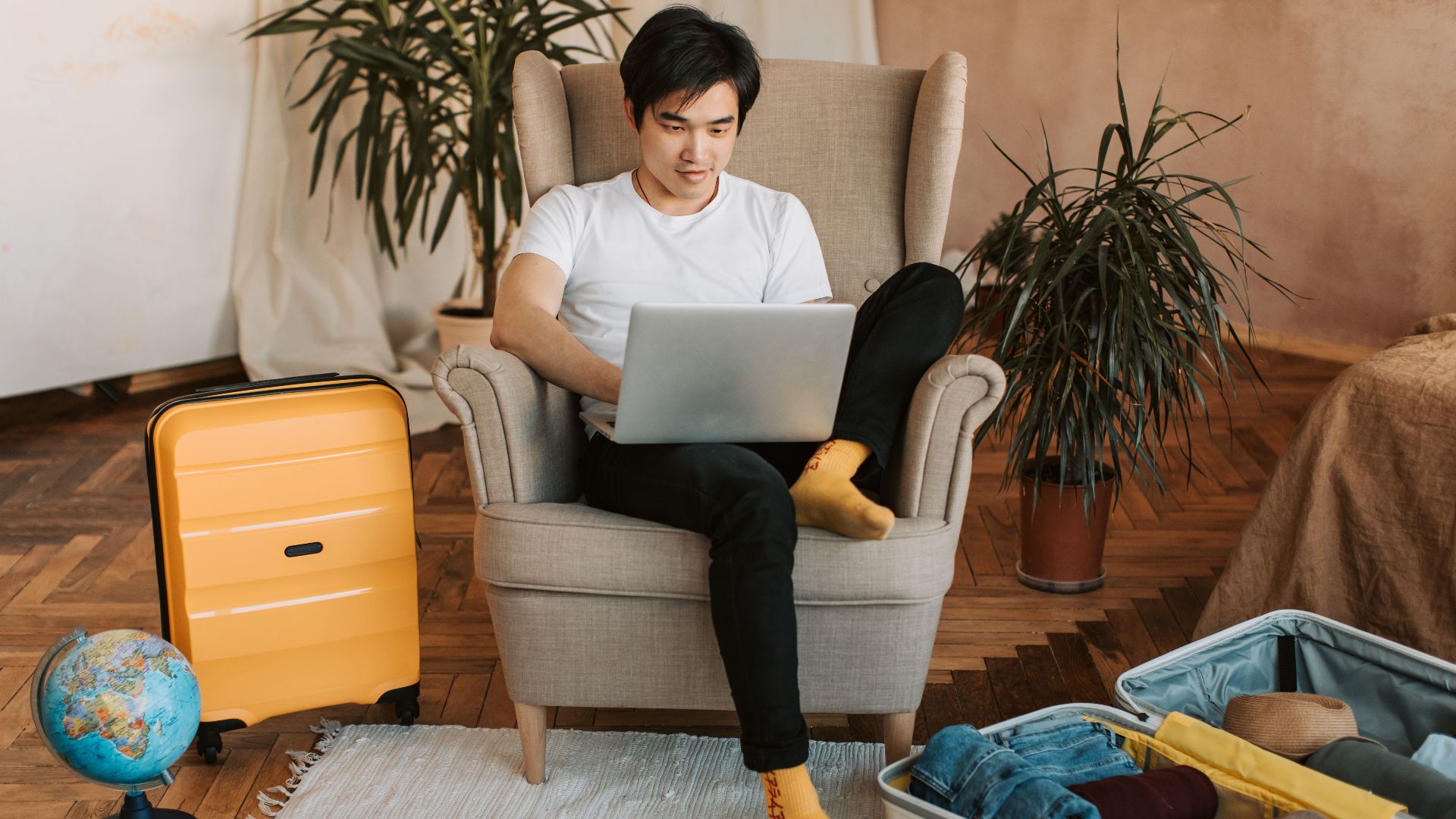 Man sitting on armchair planning travel, using laptop, with suitcases and globe nearby.