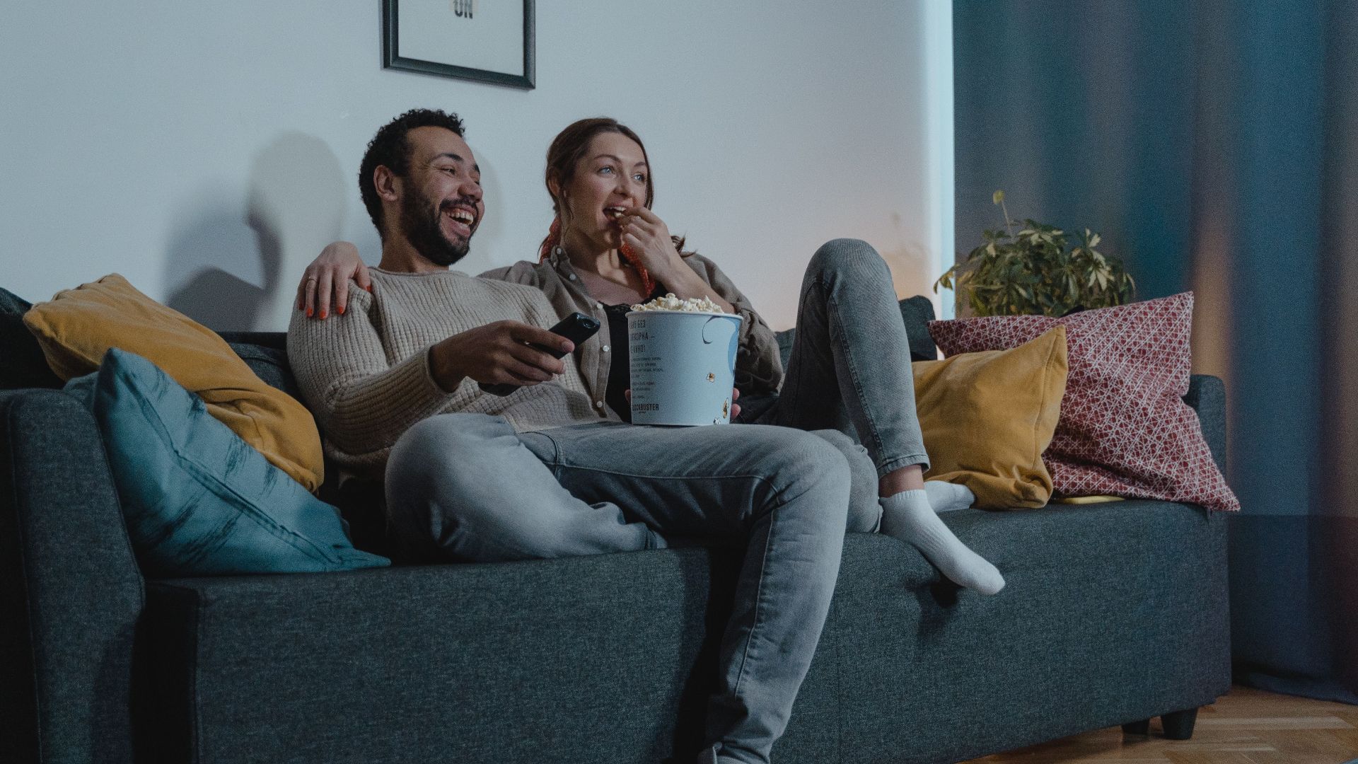 A couple relaxes indoors with popcorn, watching TV on a comfortable sofa.