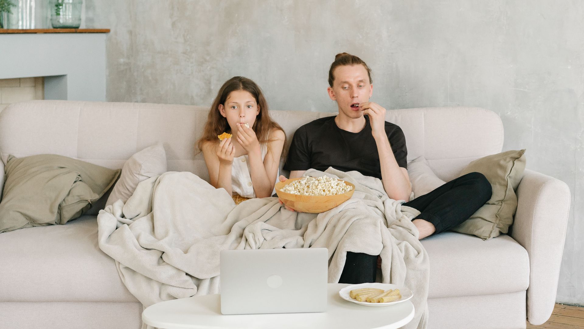 Father and daughter enjoying a cozy movie night with popcorn and snacks on a living room couch.