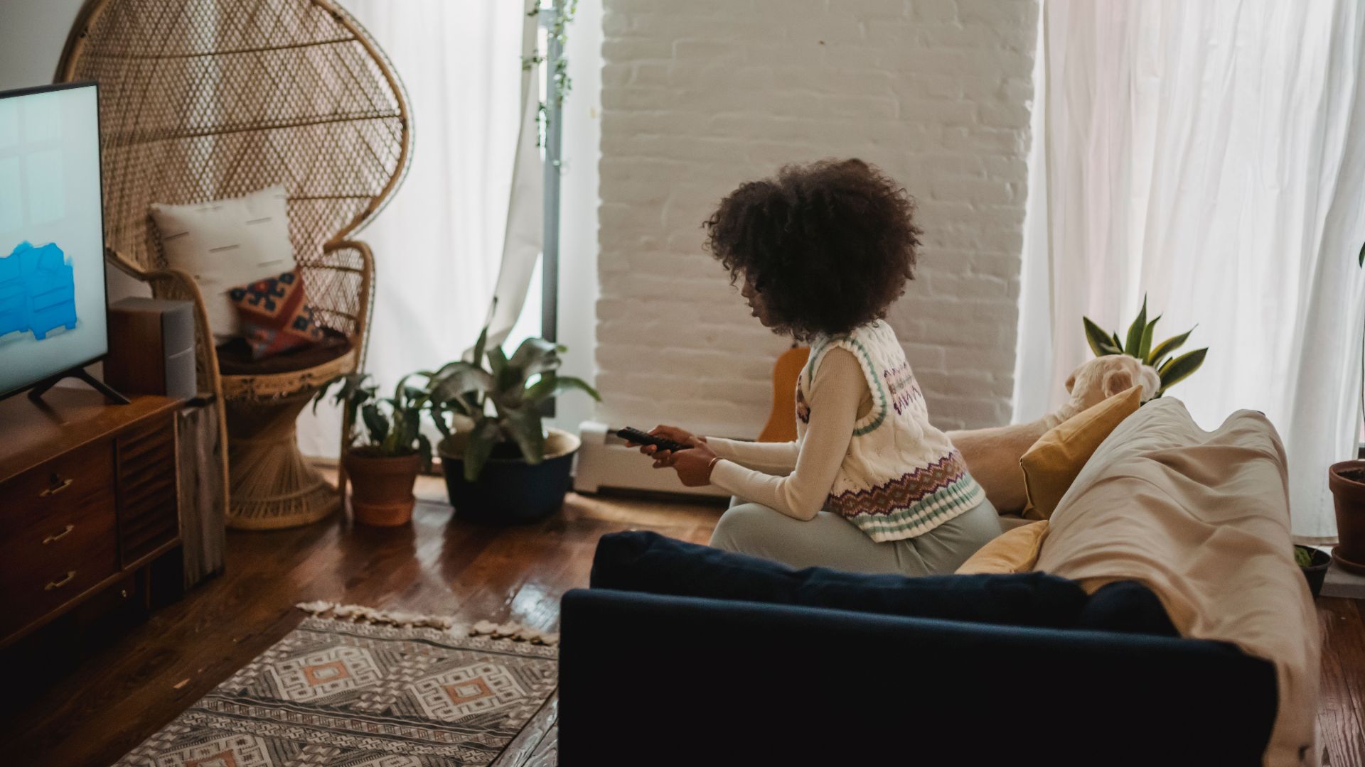 Side view of young African American female millennial with dark curly hair in stylish clothes switching channels on TV with remote controller while resting on sofa during weekend at home