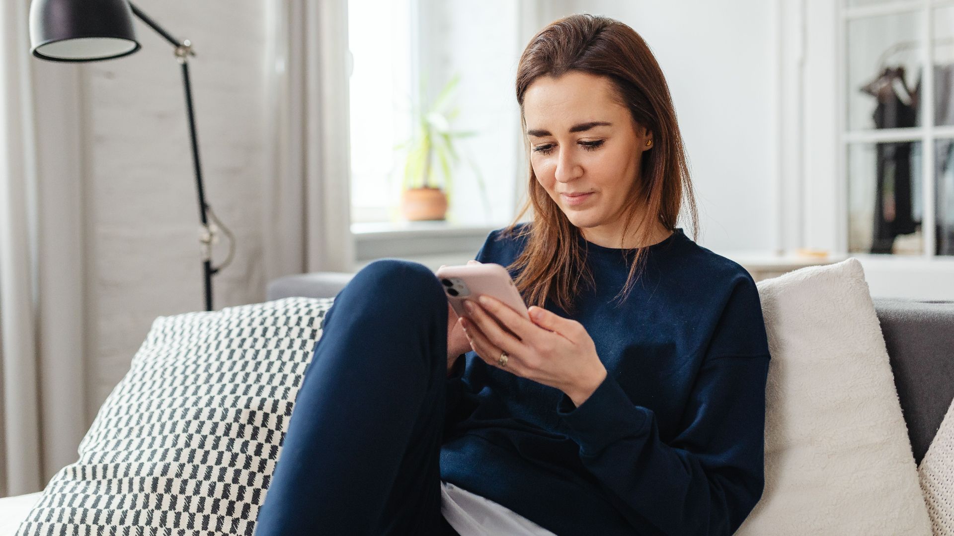 Woman in a blue sweater using a smartphone indoors, relaxing on the couch.