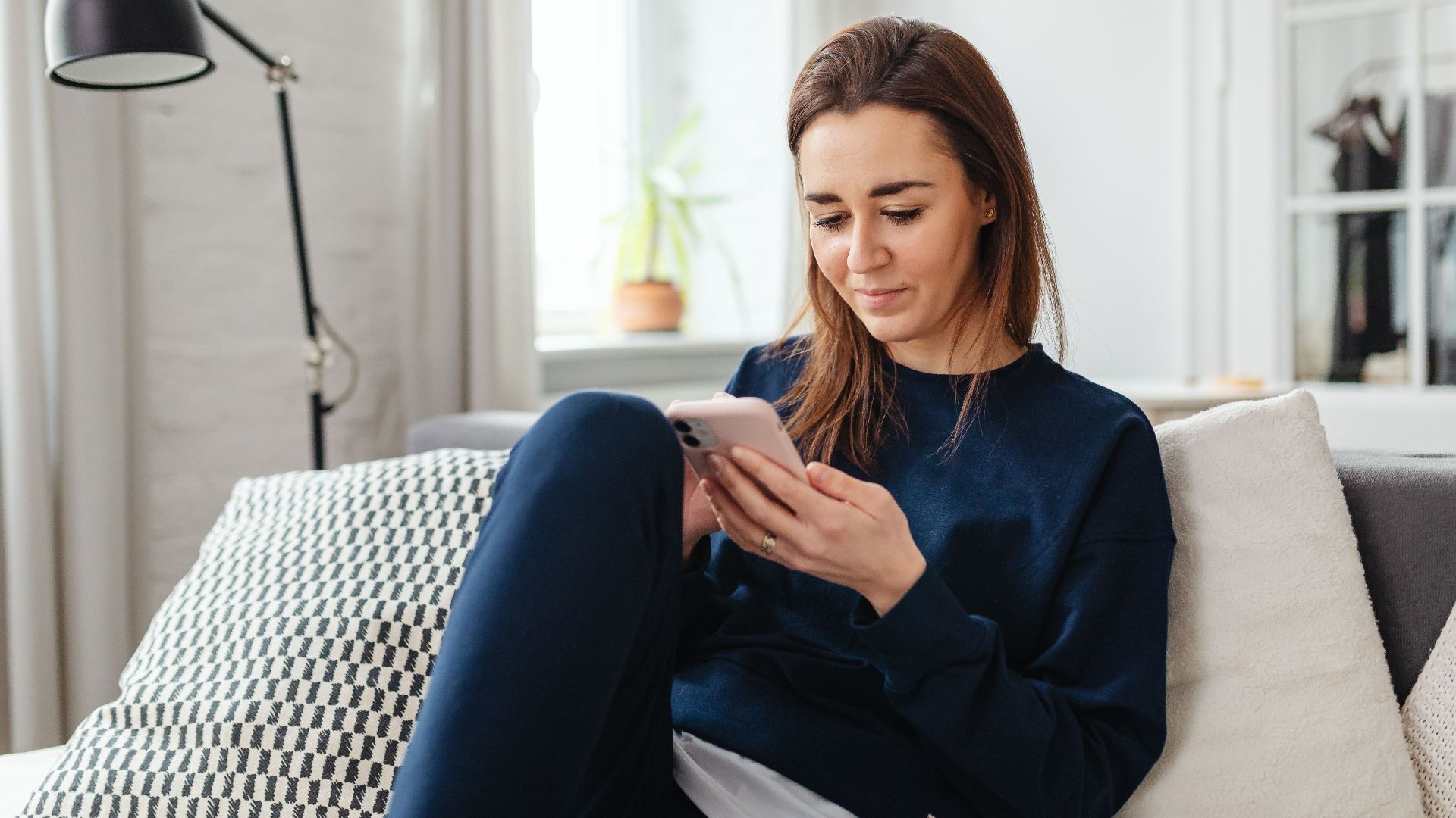Woman in a blue sweater using a smartphone indoors, relaxing on the couch.