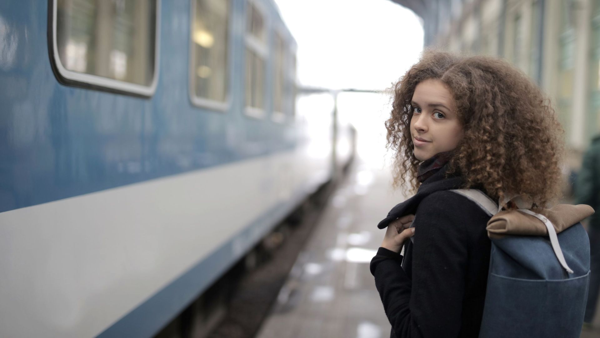 Back view of young student with curly hair in casual warm coat standing on platform with backpack near train and looking at camera over shoulder
