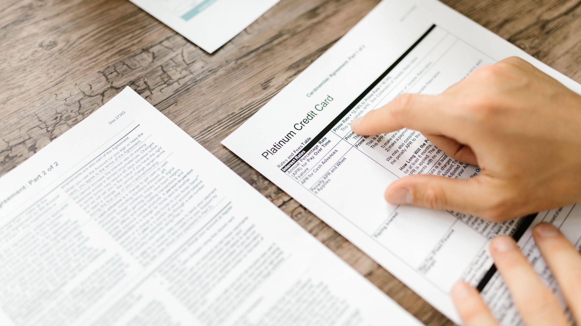 A hand examining a credit card agreement on a wooden desk, highlighting financial review.