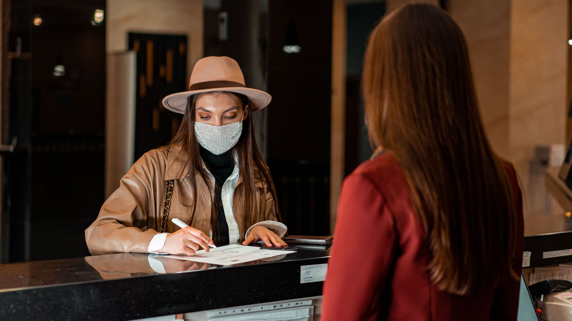 A woman in a hat and face mask checks in at a hotel reception, interacting with a receptionist.
