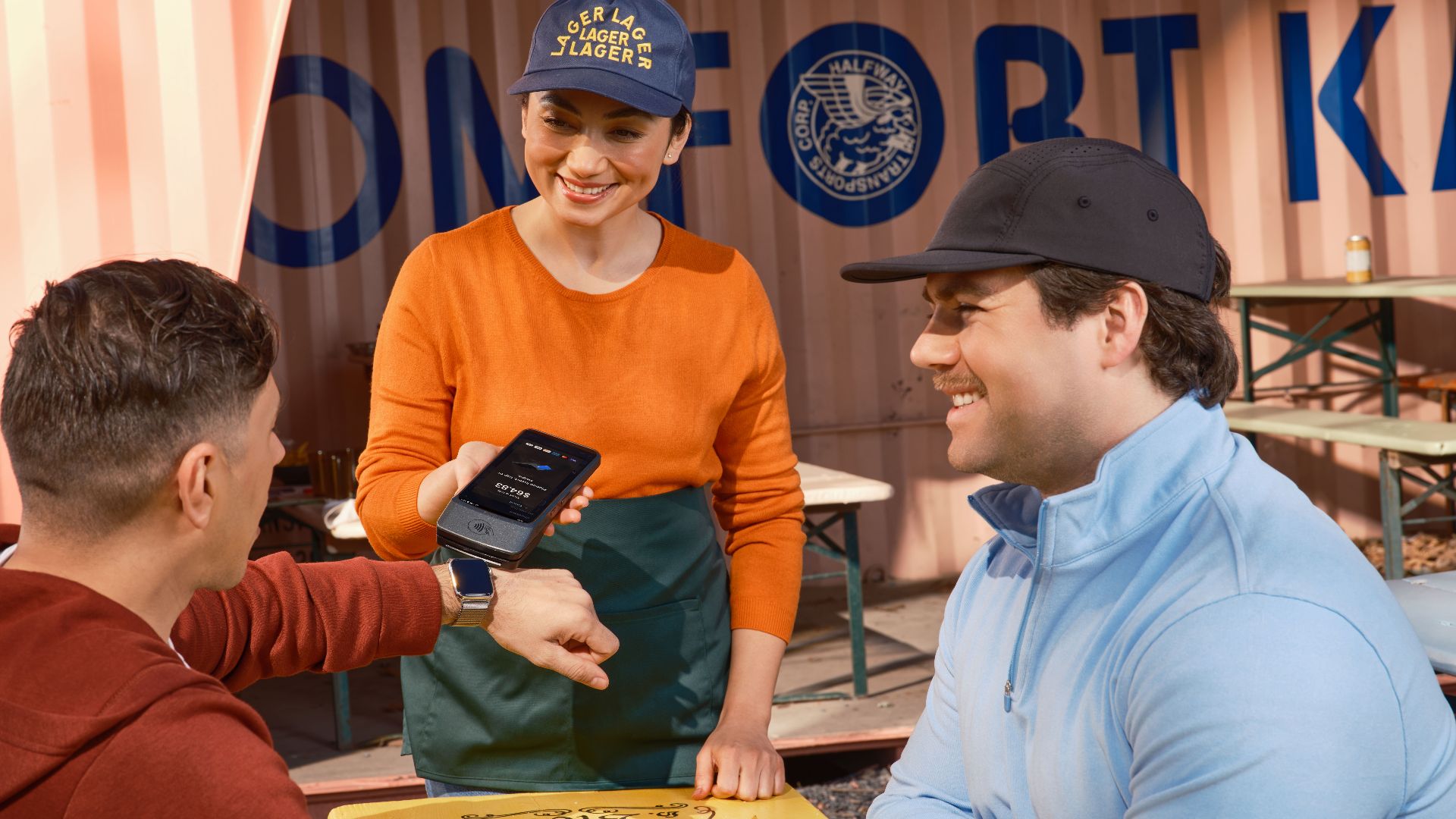 Friends using smartwatch for contactless payment at an outdoor cafe, assisted by a smiling waitress.