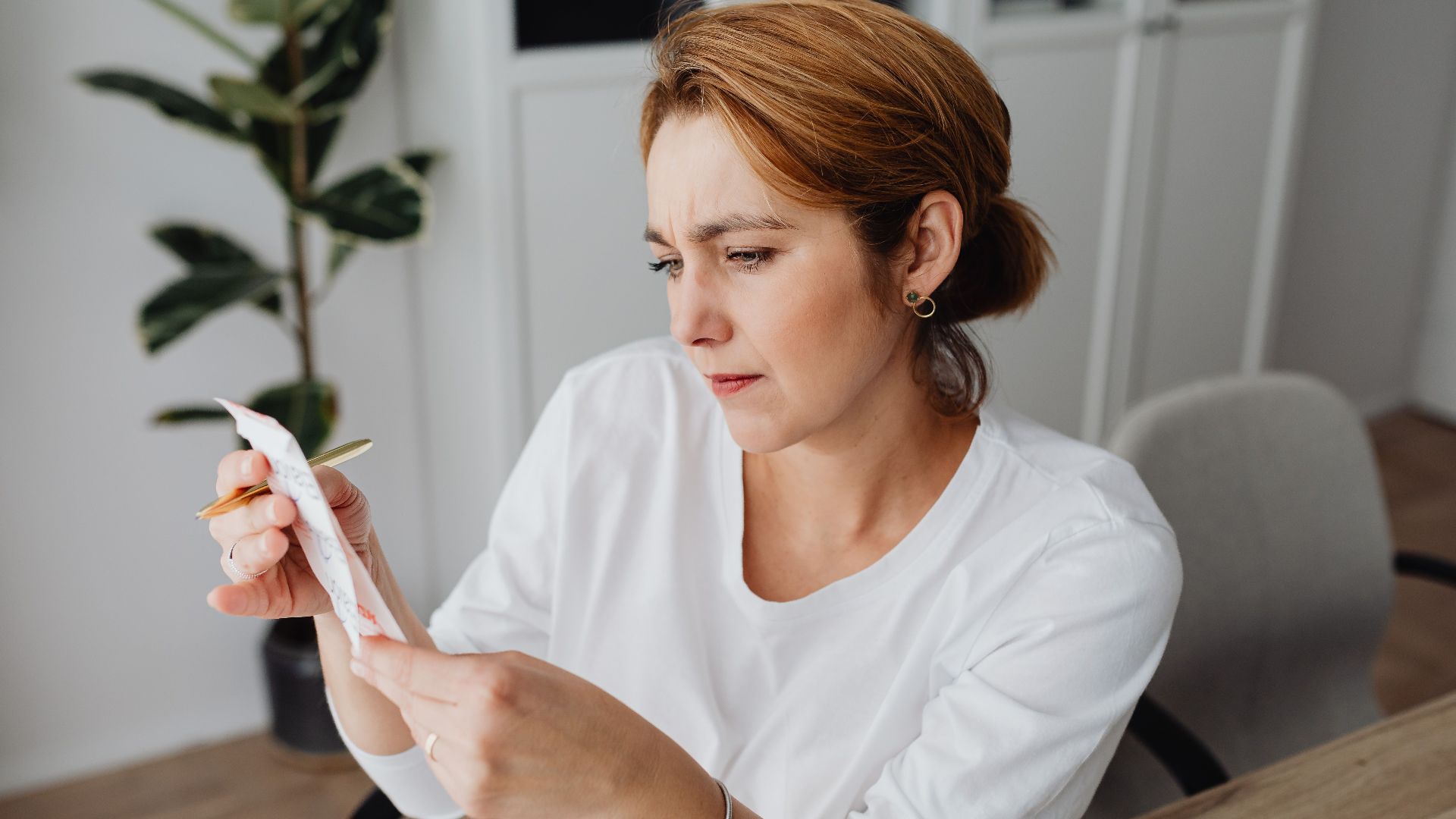 A concerned adult woman examines a receipt while sitting at her office desk, indicating financial scrutiny.