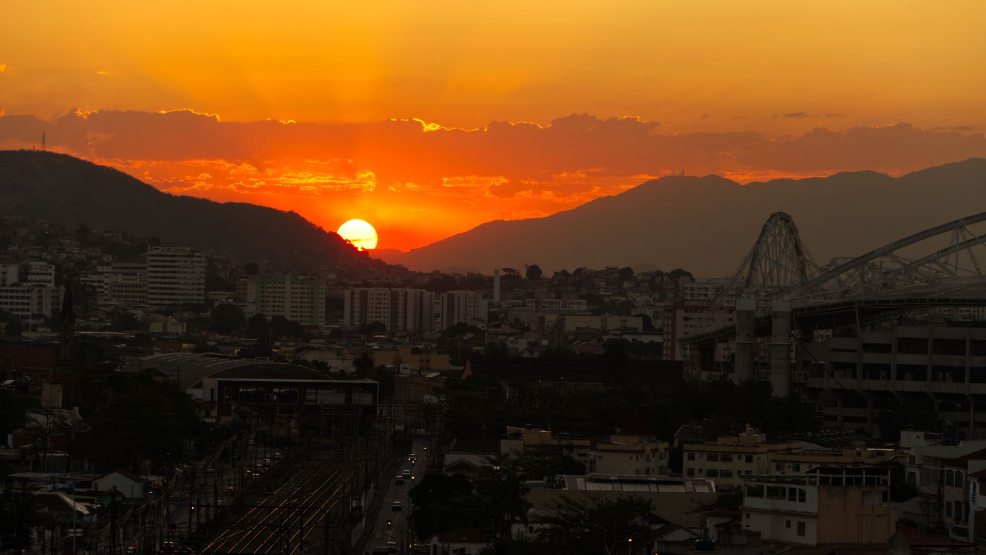 Vivid sunset over a city with mountainous backdrop and bridge silhouette.