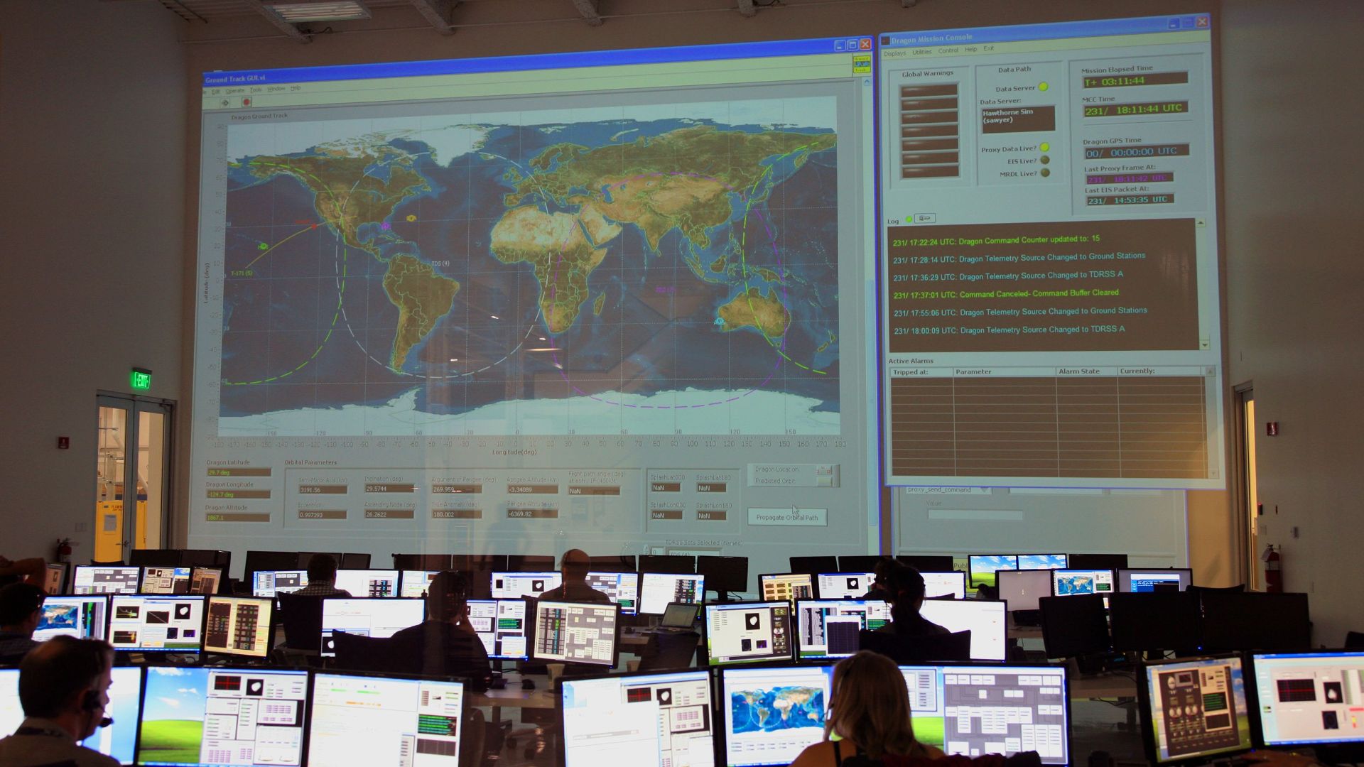 Back view of unrecognizable employees working on computers in flight control room with big interactive map on wall and analyzing data