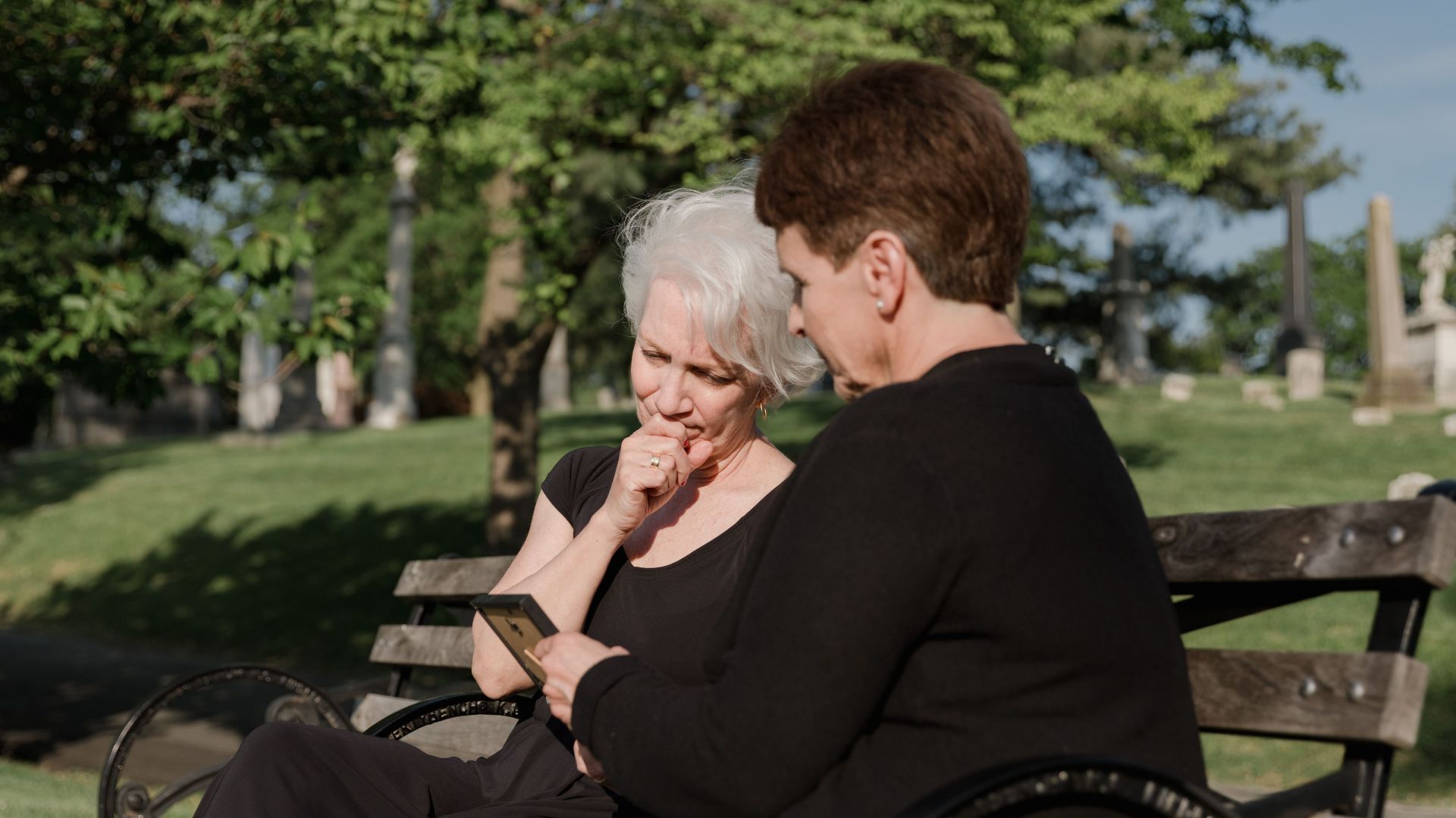 Two senior women share a moment of grief, sitting on a cemetery bench, holding a picture frame.