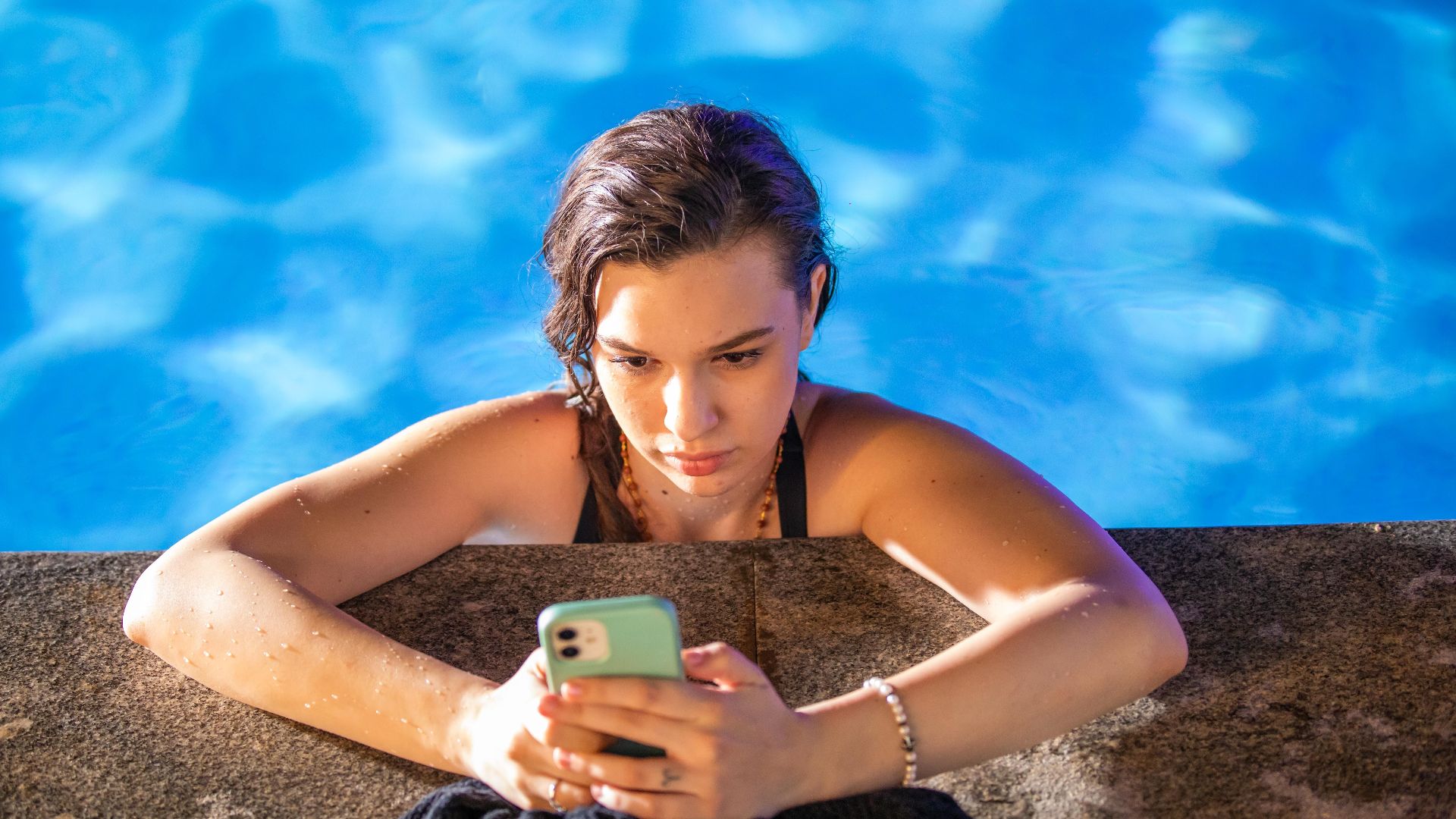 A young woman in a pool checks her smartphone under the bright sun.