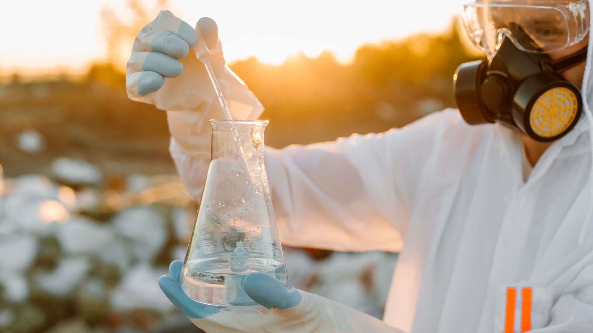 Researcher in protective suit with Erlenmeyer flask outdoors at sunset.