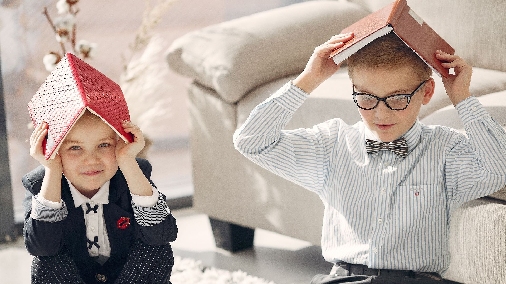 Smiling little students in formal clothes gathering on floor near sofa and playing games with textbooks while studying together at home