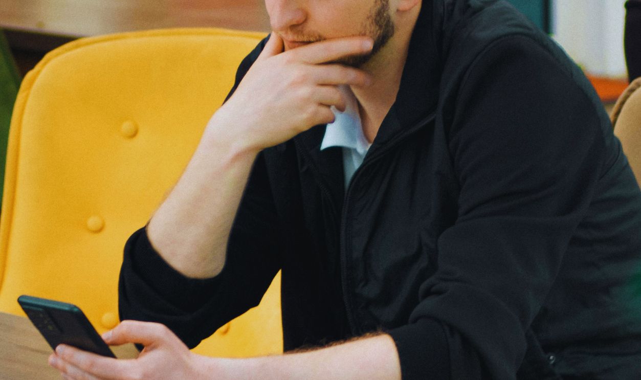 A man in a baseball cap sits indoors, thoughtfully using his smartphone at a table.