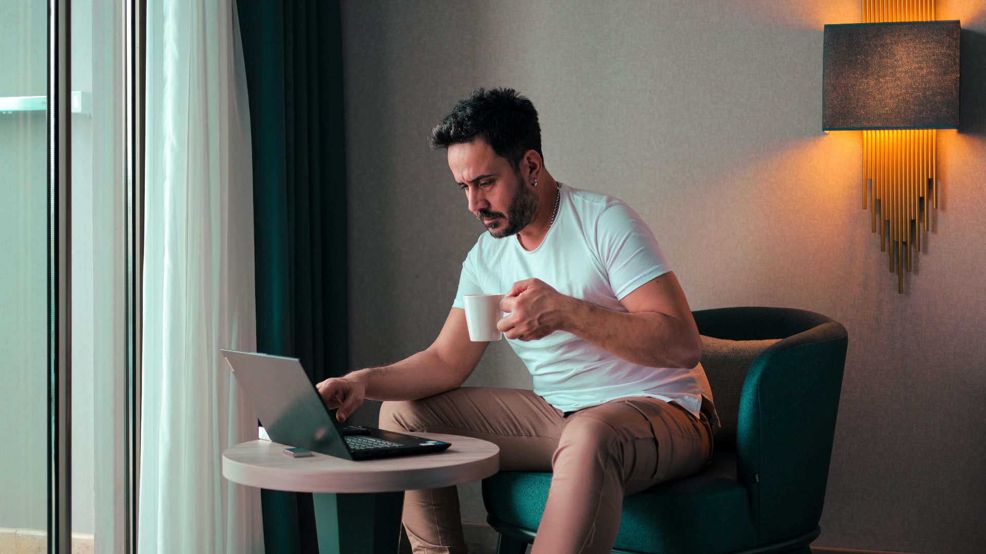 A man sits in a cozy hotel room in Cyprus, working on a laptop while enjoying a coffee.