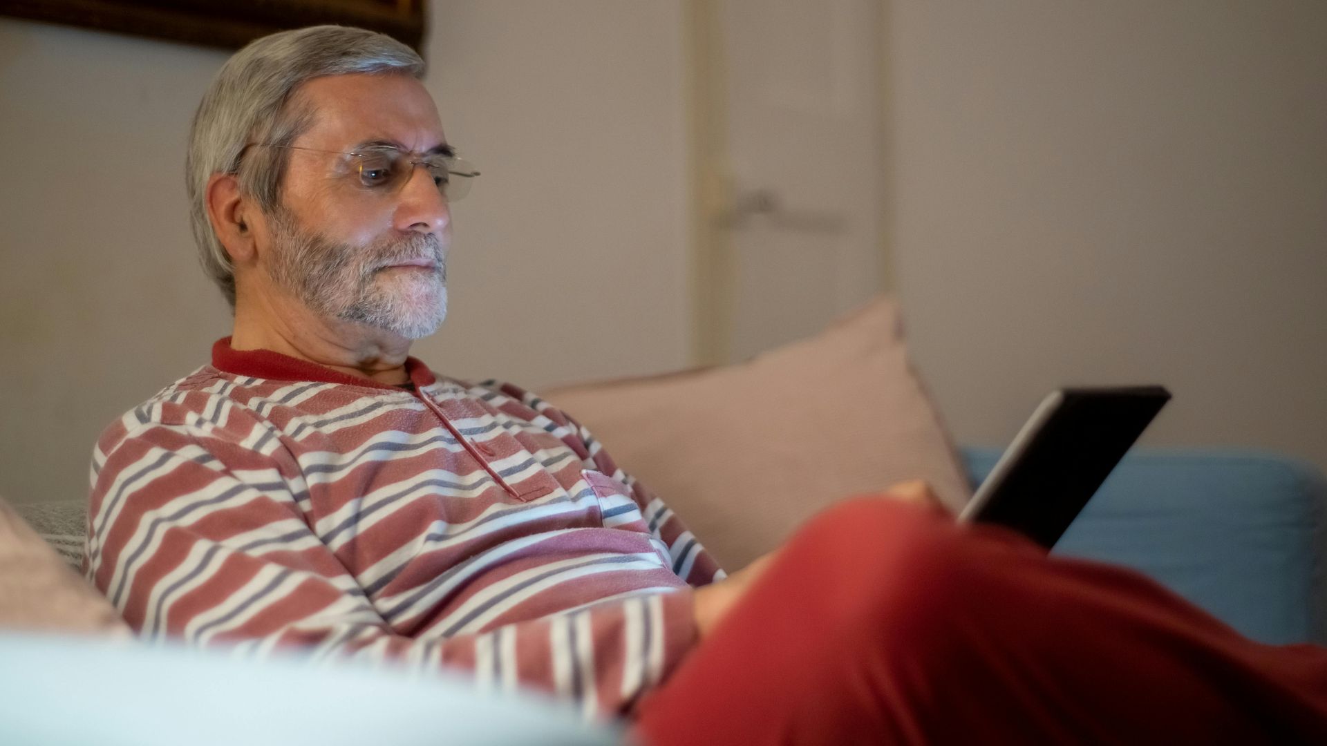 Senior man in glasses with a striped shirt using a tablet at home.