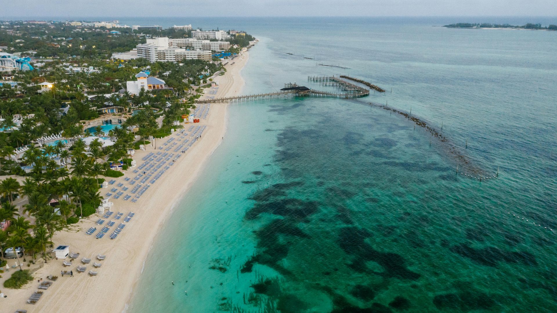 A tranquil aerial view of a beachfront resort, white sandy beach, and an extensive pier stretching into the turquoise sea.