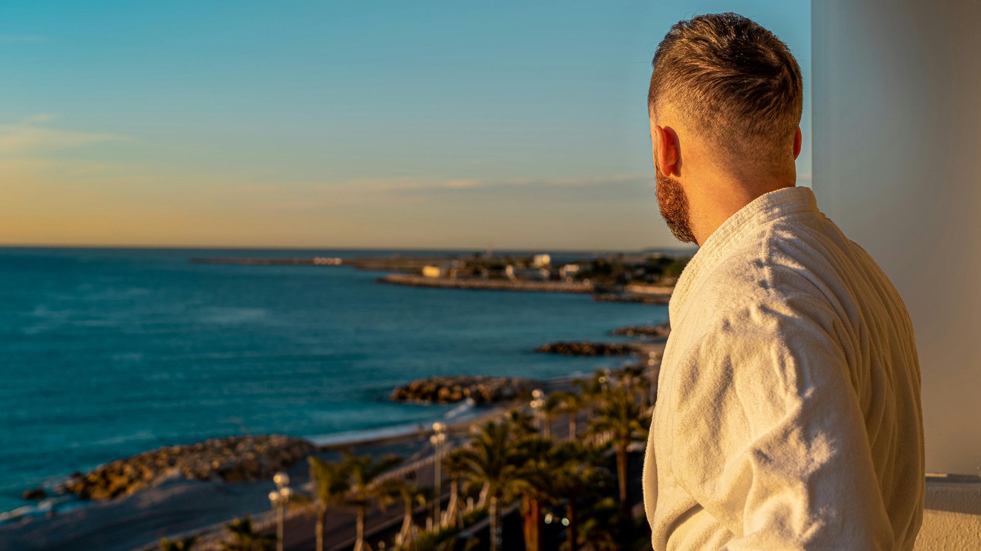 Man in bathrobe enjoys tranquil coastal view from balcony in Nice, France during sunrise.