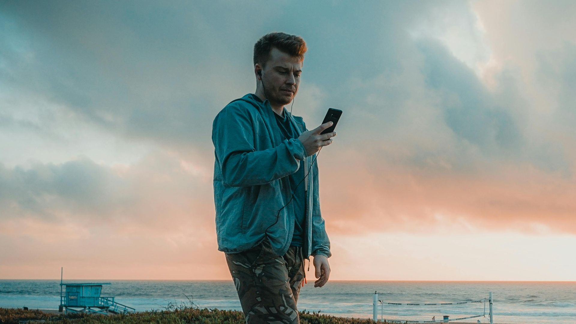A man rollerblades along a beachside path, using a smartphone under a vibrant sunset sky.
