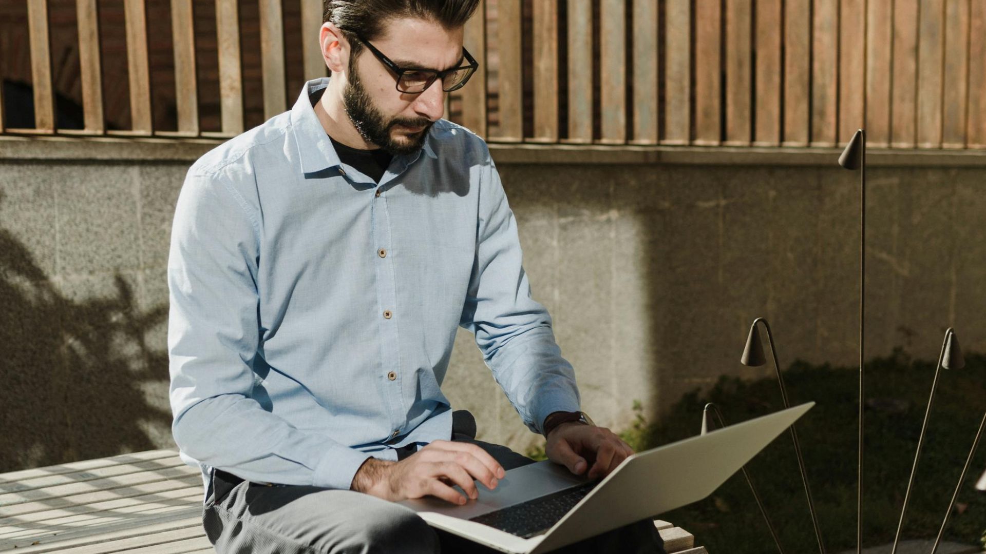 Caucasian man sitting outdoors working on laptop in sunlight with casual attire and eyeglasses.